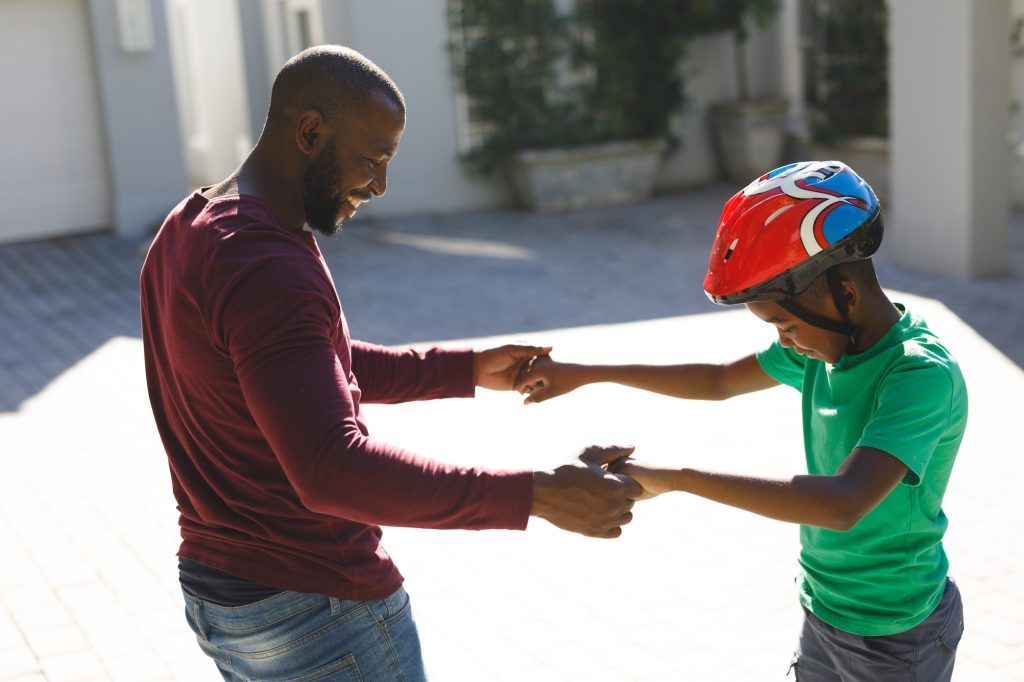 African american father smiling and helping son balancing on skateboard in garden