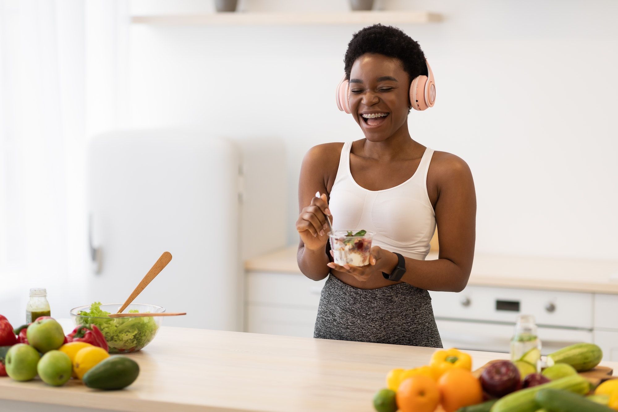 African Lady Eating Healthy Dessert Standing In Kitchen, Wearing Earphones